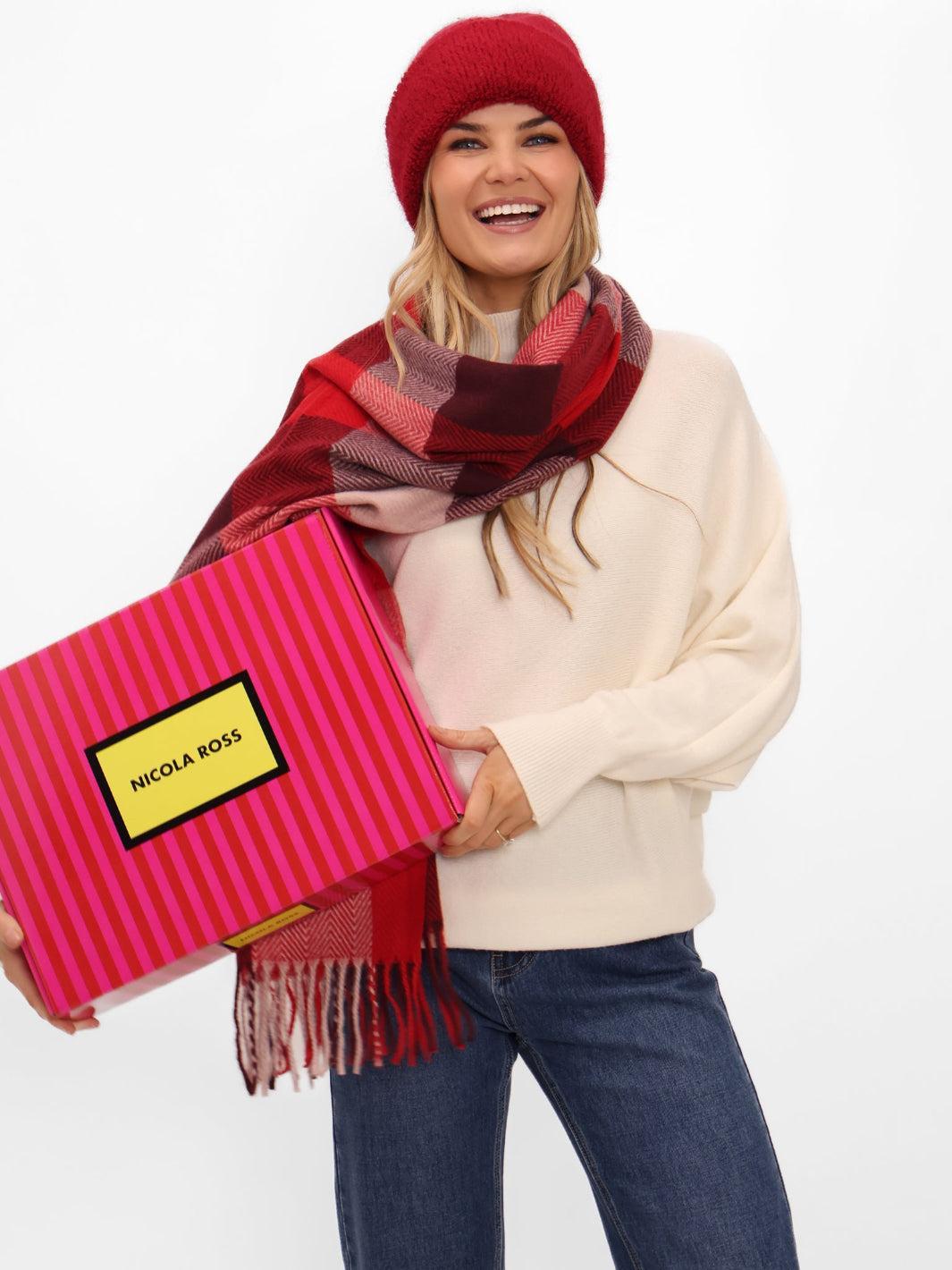 Woman holding a pink gift box with a red and pink plaid scarf, wearing a red beanie and white sweater.