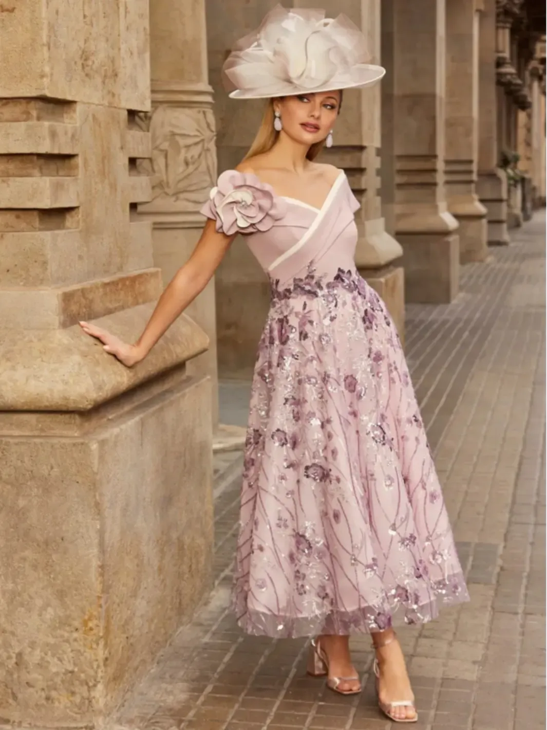Woman in a floral dress and hat standing against a stone building.