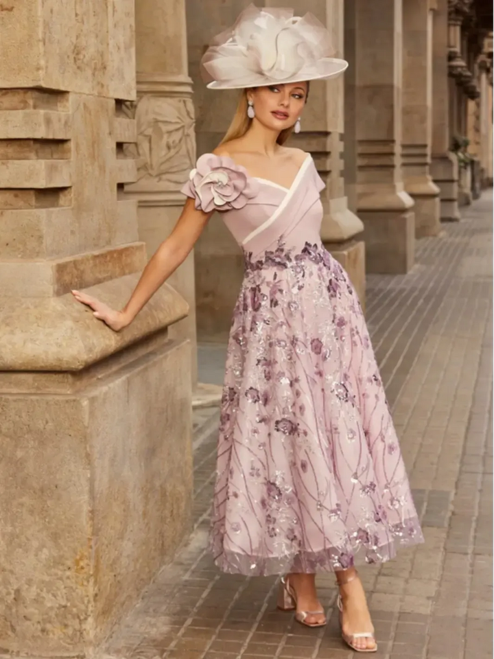 Woman in a floral dress and hat standing against a stone building.