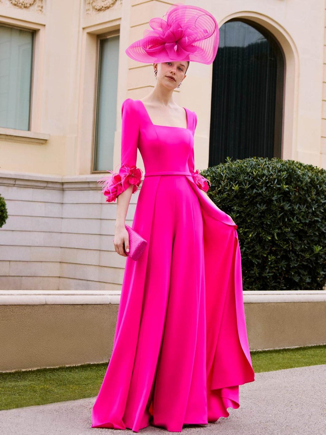 Woman in a bright pink gown and matching hat standing in front of a building.