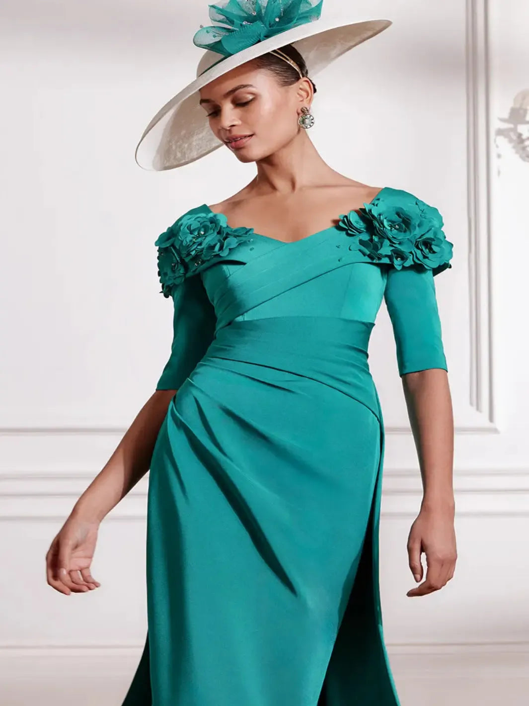 Woman wearing a teal dress with floral embellishments and a matching hat indoors.