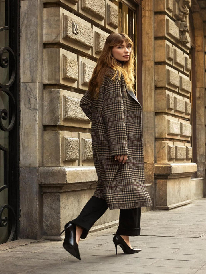 Woman in a long coat walking along a stone-paved street with ornate building facades.