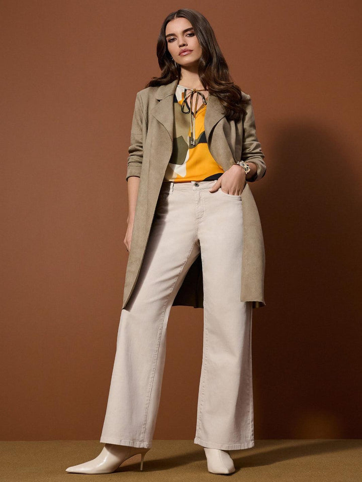 Woman wearing a beige coat, yellow top, and white pants against a brown background