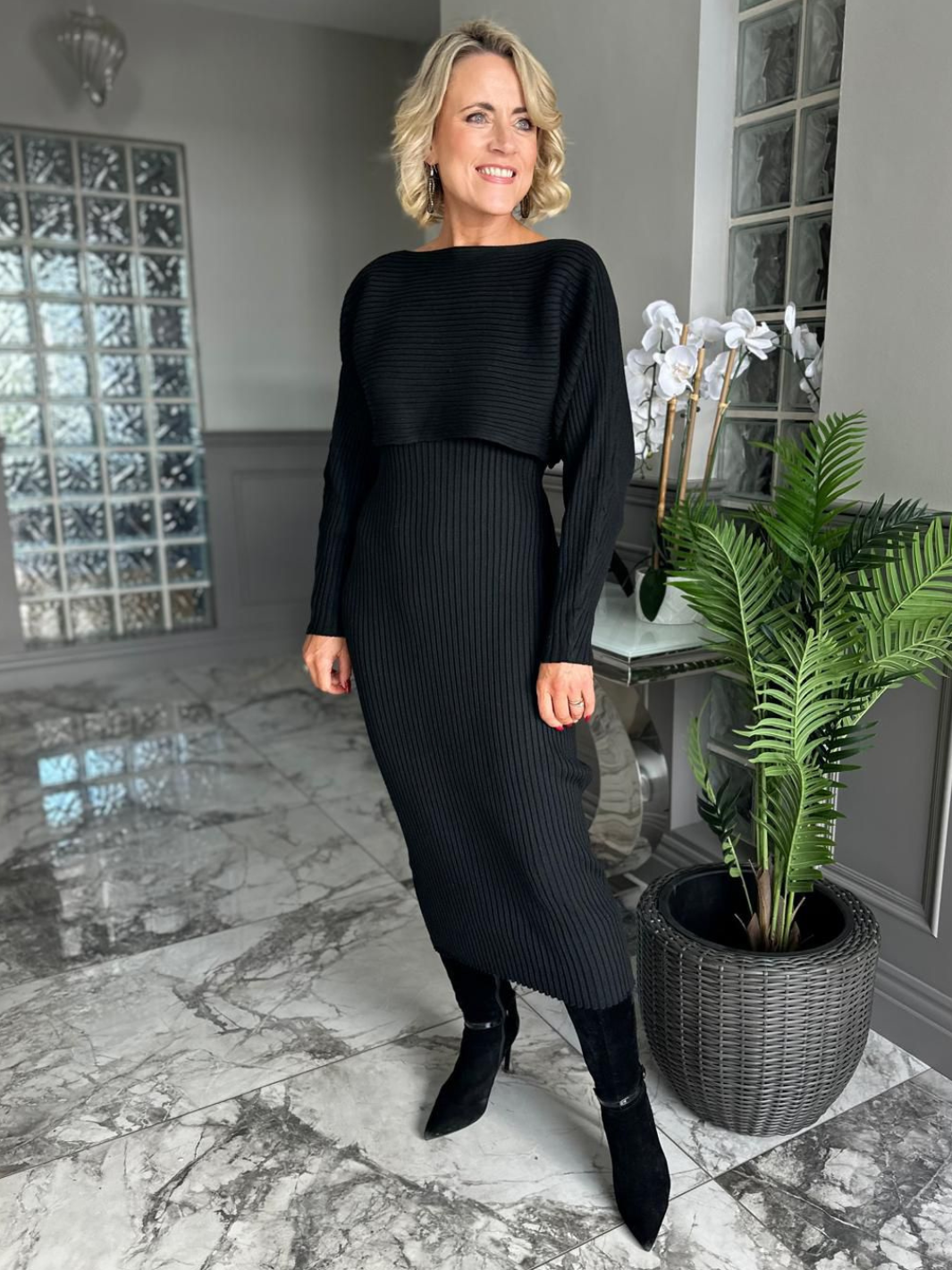 Woman in a black outfit standing in a modern interior setting with a plant.