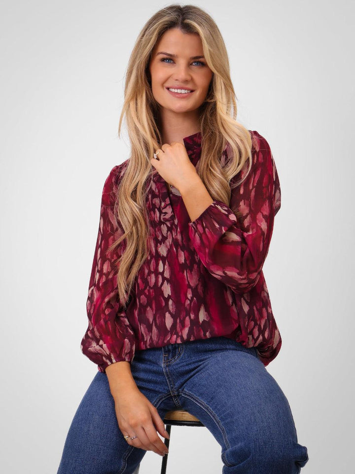 Woman wearing a patterned blouse and jeans sitting on a stool against a white background