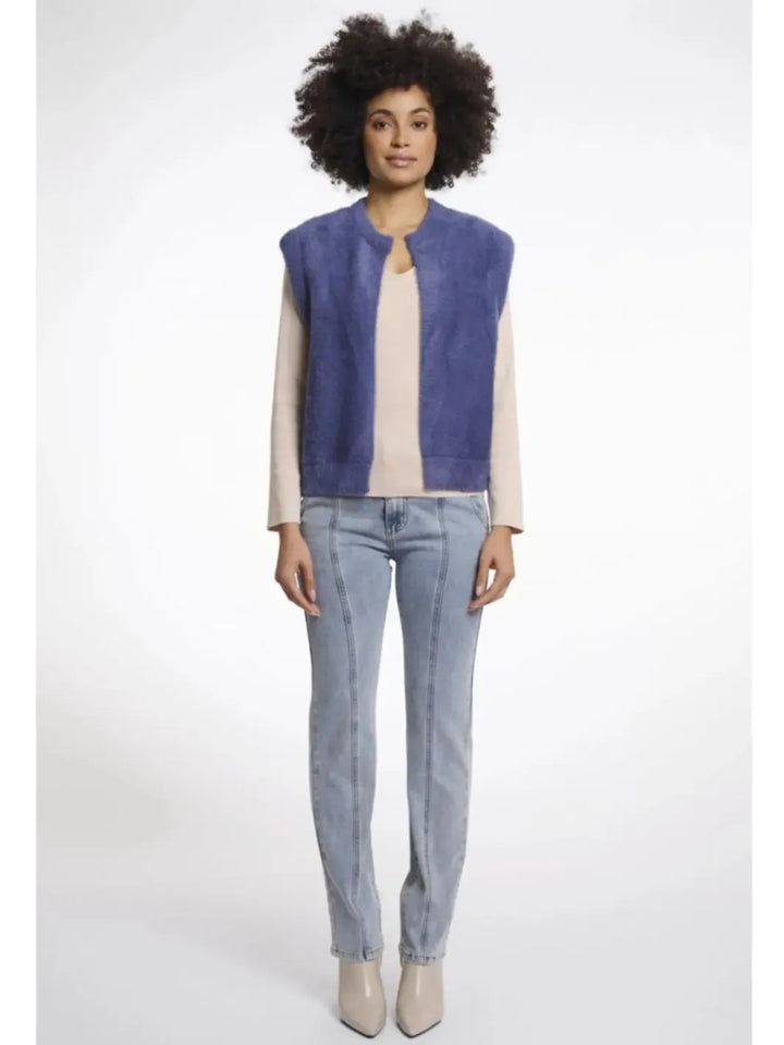 Person wearing a blue vest over a beige top and light blue jeans on a white background