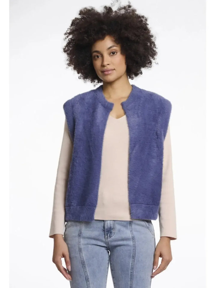 Woman wearing a blue vest over a beige shirt with light blue jeans on a white background