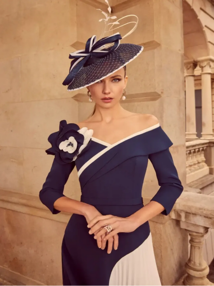 Woman wearing a navy and white dress with a matching hat, standing against a stone wall.